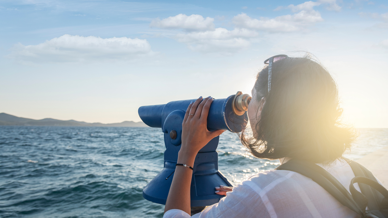  Young woman looks through a telescope by the sea on a bright day. 
